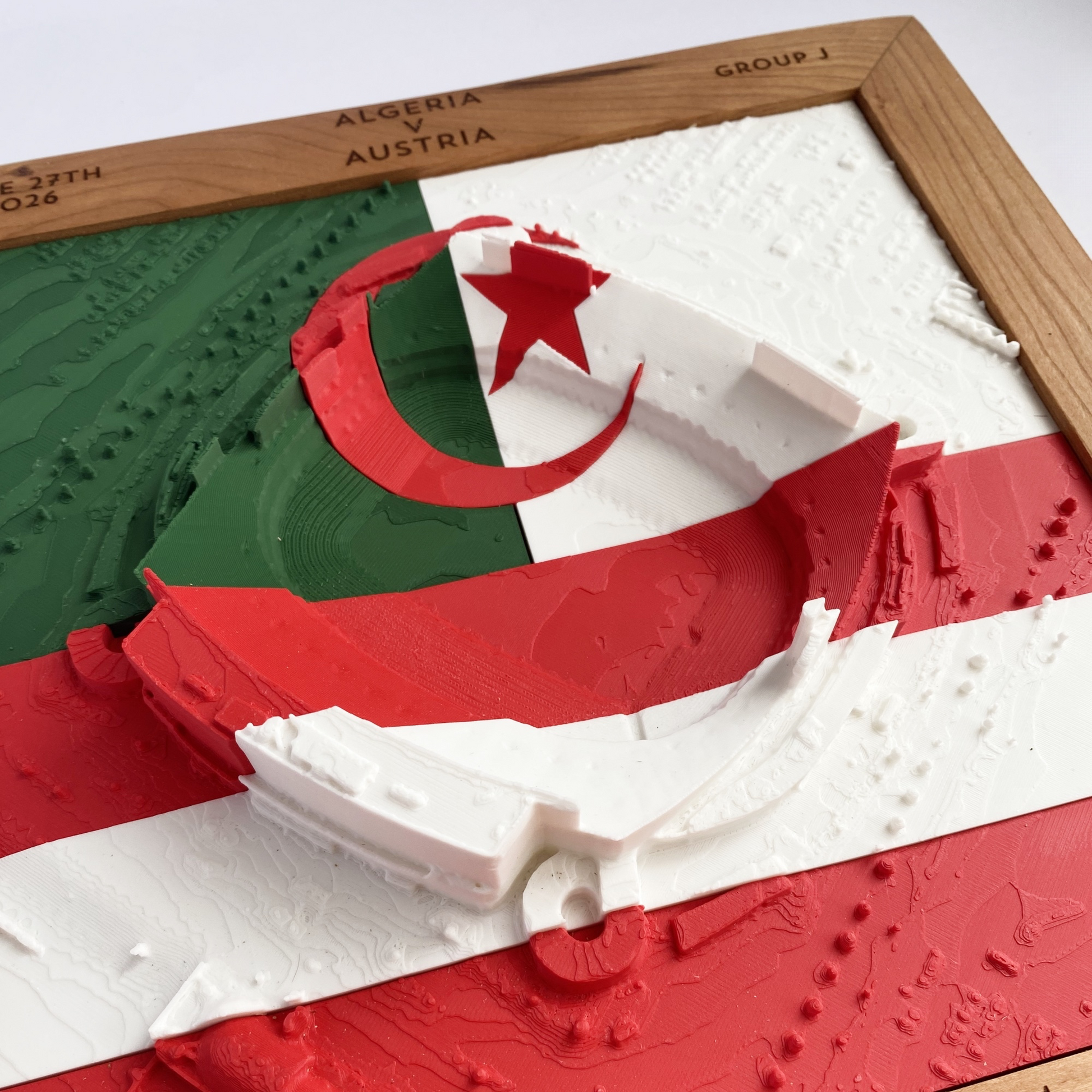 3D printed model of Arrowhead Stadium imprinted with Algeria's flag (solid green, solid white, with red moon and star) on the top half and Austria's flag (two red stripes with white stripe in between) on the bottom half - encased in a solid wooden frame engraved with the text "June 27th 2026; Algeria v Austria; Group J; Kansas City Stadium, Kansas City, MO, and coordinates"